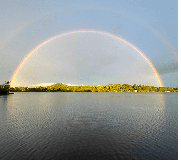 A full double rainbow arches over a calm lake with greenery.