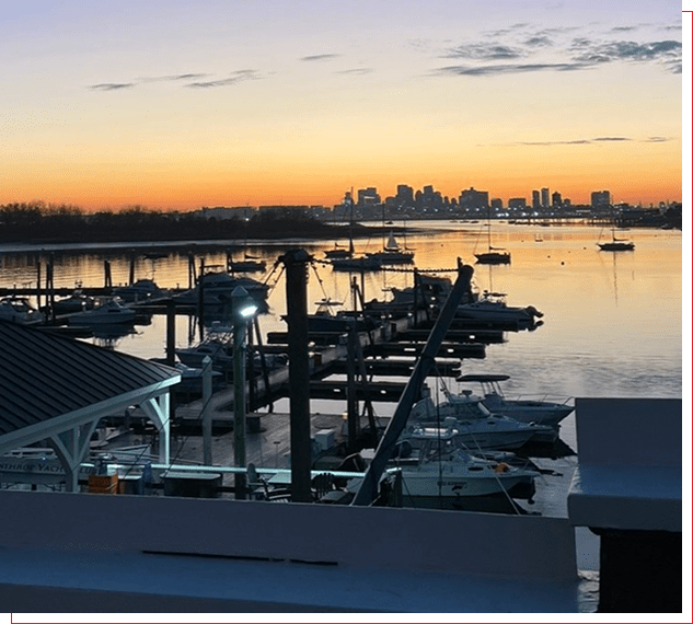 Sunset over a marina with boats and city skyline in the background.