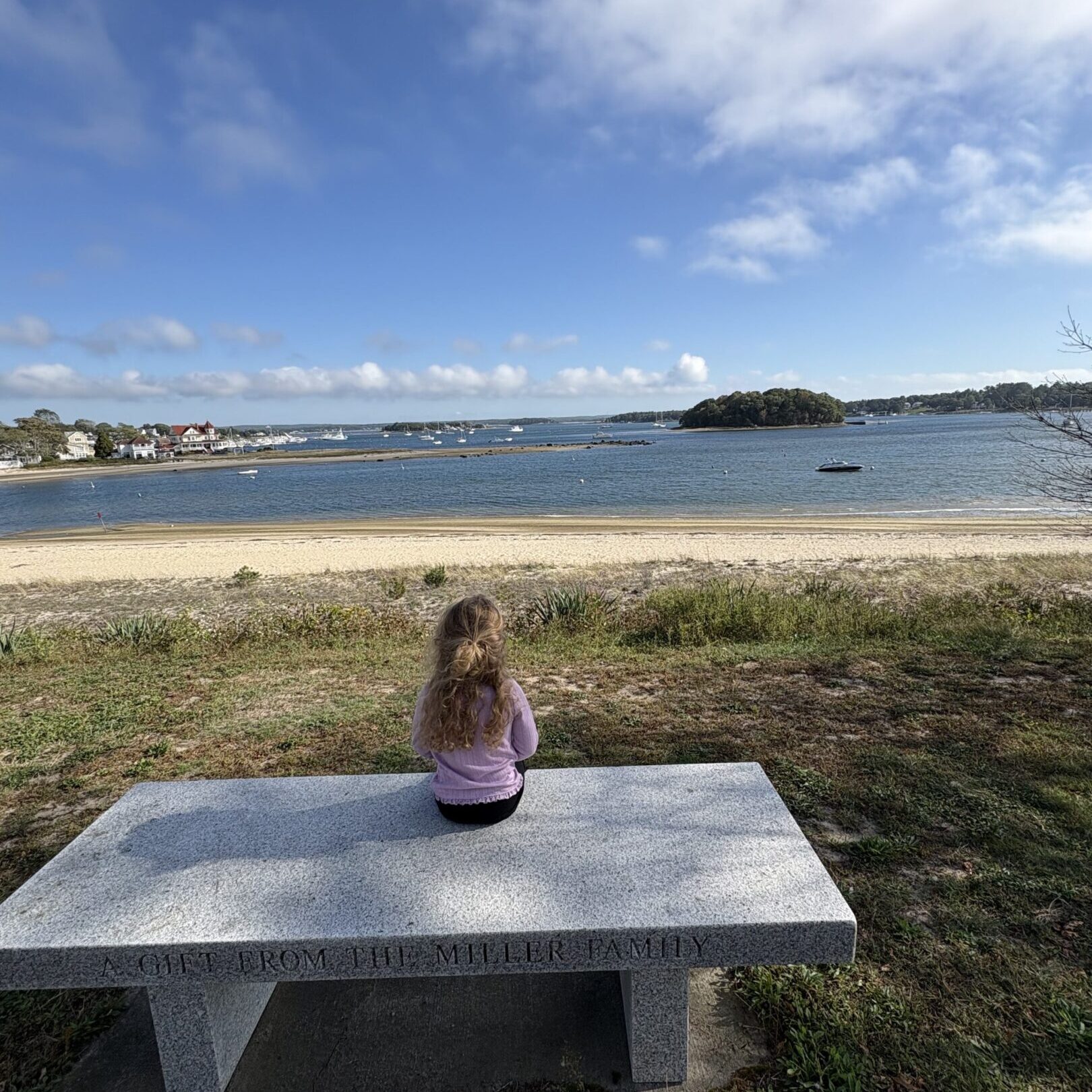 A child sitting alone on a bench by the beach under a blue sky.
